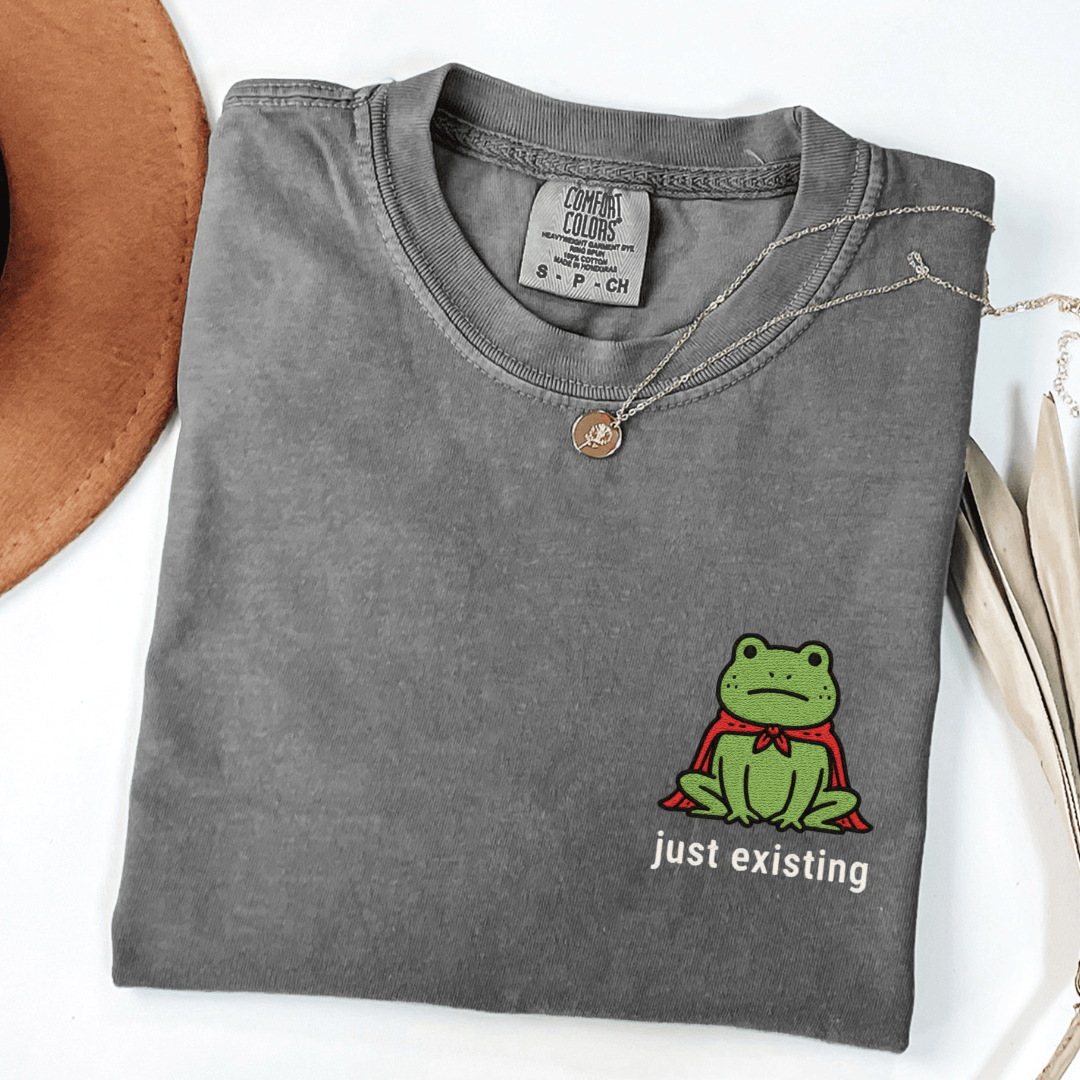 Folded Comfort Colors 1717 t-shirt in the color pepper with an embroidered frog graphic and 'just existing' text underneath the frog.